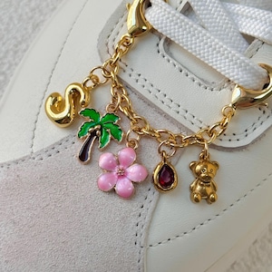 May include: Gold-toned shoe charm with a chain holding various charms, including a letter "S", a palm tree, a pink flower, a teardrop-shaped gem, and a teddy bear. The charm is attached to a white sneaker with white laces.