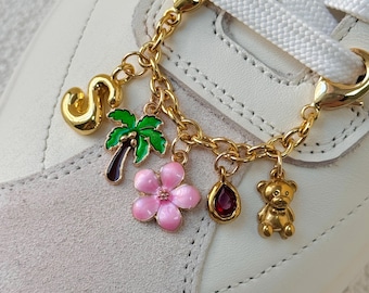 May include: Gold-toned shoe charm with a chain holding various charms, including a letter "S", a palm tree, a pink flower, a teardrop-shaped gem, and a teddy bear. The charm is attached to a white sneaker with white laces.