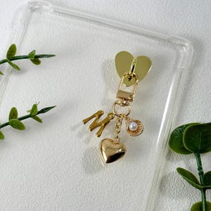 Pode incluir: Chaveiro dourado com um pingente em forma de coração, a letra "M", um pequeno coração e uma concha com uma pérola. O chaveiro tem um fecho de lagosta e um topo em forma de coração. O fundo é uma capa de telefone transparente.
