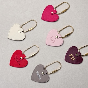 A flat lay of six heart-shaped leather keychains in elegant colors including red, pink, burgundy, beige, grey, and fuchsia. Each keychain features a gold metal clasp and personalized details such as initials, names, or embossed letters in different finishes like gold foil, silver foil, and engraving. The soft leather texture and minimalist design create a stylish and romantic aesthetic.