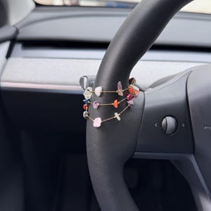 Könnte beinhalten: Ein mehrreihiges Armband mit bunten, unregelmäßig geformten Edelsteinen. Das Armband ist über einem schwarzen Lenkrad drapiert. Die Edelsteine umfassen rosa, lila, orange und grüne Steine, die auf zarten Goldketten aufgefädelt sind.