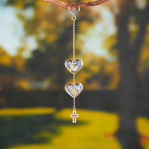 Könnte beinhalten: Goldfarbenes Hängeornament mit zwei herzförmigen Anhängern, gefüllt mit blauen und weißen Blumen. Unter den Herzen hängt ein kleines goldenes Kreuz. Das Ornament wird von einer Kette an einem Ast aufgehängt.