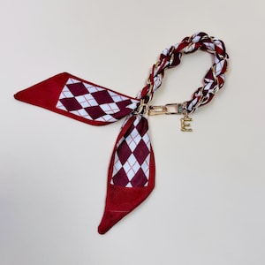 Puede incluir: Un pa&ntilde;uelo con estampado burdeos y blanco con una cadena y cierre dorados. El pa&ntilde;uelo presenta un patr&oacute;n de rombos y un charm con la letra "E" dorada. La cadena est&aacute; trenzada con tela burdeos y blanca.
