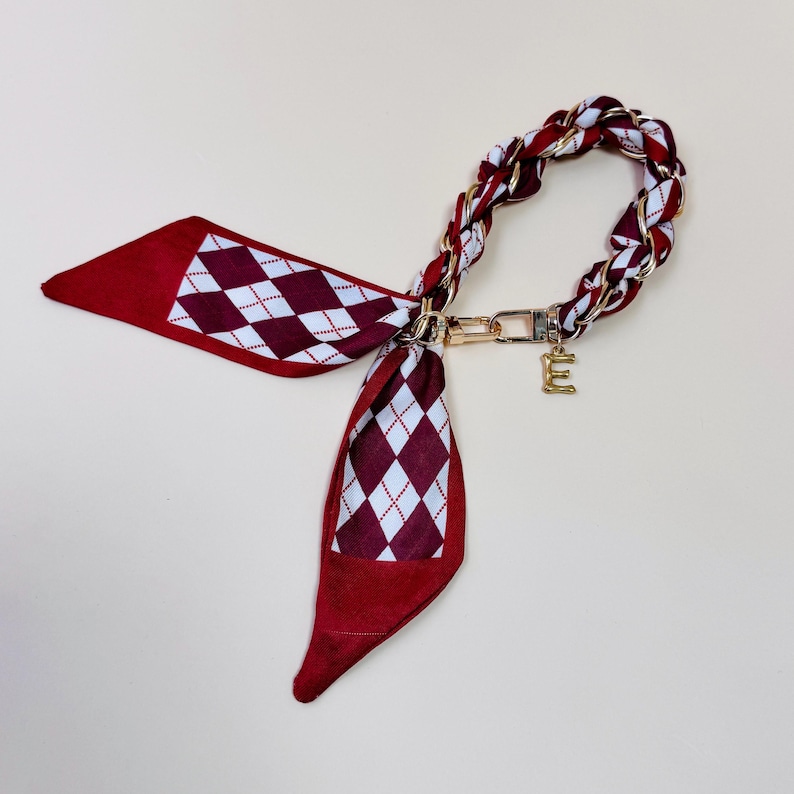 Puede incluir: Un pa&ntilde;uelo con estampado burdeos y blanco con una cadena y cierre dorados. El pa&ntilde;uelo presenta un patr&oacute;n de rombos y un charm con la letra "E" dorada. La cadena est&aacute; trenzada con tela burdeos y blanca.