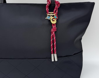 May include: Black tote bag with a quilted bottom and black handles. A decorative red and white rope charm with gold accents and a gold letter "G" hangs from the handle.