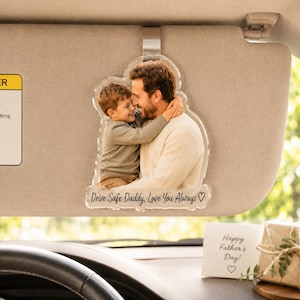 Peut inclure: Un clip de pare-soleil en acrylique transparent avec une photographie imprimée d'un enfant embrassant un adulte. Le texte "Drive Safe Daddy, Love You Always" accompagné d'un cœur est imprimé en noir sur le bas de la plaque en acrylique.