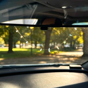 Könnte beinhalten: Eine zarte goldene Halskette mit kleinen, unregelmäßig geformten, hellblauen und weißen Edelsteinen. Die Halskette hängt vor der Windschutzscheibe eines Autos, mit einem verschwommenen Hintergrund aus Bäumen und Sonnenlicht.