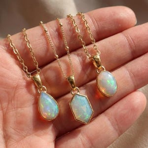 May include: Three gold necklaces with opal pendants in teardrop, hexagonal, and oval shapes. The opals display iridescent colors. The necklaces are held in a hand, showcasing the jewelry's design and craftsmanship.