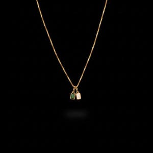 May include: A delicate gold necklace with two small pendant charms. One charm is a rectangular emerald green stone, and the other is a rectangular white stone. The necklace hangs against a black background.
