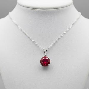 Op de afbeelding: Een zilveren ketting met een ronde, dieprode edelsteen hanger. De hanger is gezet in een zilveren zetting met een kleine, heldere accentsteen boven de robijn. De ketting wordt getoond op een witte mannequin.
