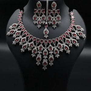 Puede incluir: Un conjunto de joyas de tres piezas que incluye un collar, pendientes y una diadema. El conjunto se exhibe en un maniquí negro. Las joyas tienen un engaste de metal plateado con detalles de piedras preciosas rojas. El collar y los pendientes tienen un diseño de lágrima.