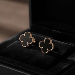 May include: A pair of stud earrings featuring a four-leaf clover shape with a smooth black center inlay and a beaded gold-toned metal border. The earrings are presented in a black velvet-lined jewelry box with a hinged lid.