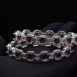 Puede incluir: Dos pulseras plateadas con piedras centrales rectangulares de color rubí y piedras de acento transparentes. Las pulseras se presentan sobre un fondo negro, resaltando el diseño y el brillo de las joyas. Las pulseras son una pieza de joyería de moda.