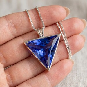 以下が含まれることがあります： 三角形のペンダントが付いたシルバーネックレス。ペンダントには、大きくファセットカットされた濃い青色の宝石がセットされています。宝石はシルバーのフレームにセットされ、繊細なチェーンから吊り下げられています。宝石のファセットが光を捉え、輝きを生み出しています。