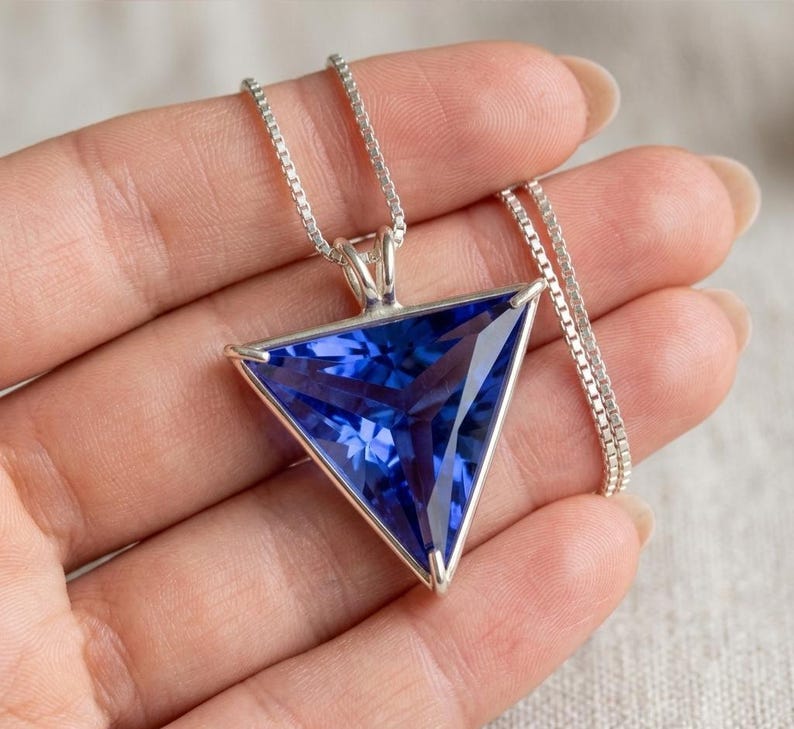 以下が含まれることがあります： 三角形のペンダントが付いたシルバーネックレス。ペンダントには、大きくファセットカットされた濃い青色の宝石がセットされています。宝石はシルバーのフレームにセットされ、繊細なチェーンから吊り下げられています。宝石のファセットが光を捉え、輝きを生み出しています。