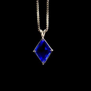 Op de afbeelding: Een zilveren ketting met een vierkante, diepblauwe hanger. De hanger heeft een levendige, juweelachtige kleur, gezet in een zilveren montuur. De ketting heeft een delicaat, geschakeld ontwerp, tegen een zwarte achtergrond.