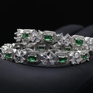 Può includere: Due braccialetti d'argento con pietre centrali quadrate verde smeraldo e dettagli chiari a forma di fiore. I braccialetti sono presentati su uno sfondo scuro, evidenziando il design intricato e i dettagli scintillanti.