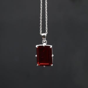 Puede incluir: Un collar de plata con un colgante rectangular de piedra preciosa rojo oscuro. El colgante está engastado en un marco de plata y cuelga de una delicada cadena de plata. El fondo es gris oscuro liso.