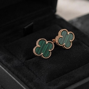 May include: A pair of stud earrings featuring a four-leaf clover shape with green malachite stone inlays and a beaded rose gold metal border. The earrings are presented in a black velvet-lined jewelry box with a structured square design.