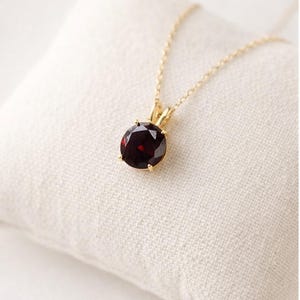 Könnte beinhalten: Eine goldene Halskette mit Anhänger, die einen runden, tiefroten Edelstein zeigt. Der Edelstein ist in einer goldenen Fassung gefasst und hängt an einer zarten Goldkette. Die Halskette wird auf einer weißen, strukturierten Oberfläche präsentiert.