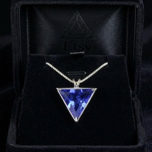 以下が含まれることがあります： 三角形の青い宝石のペンダントが付いたシルバーネックレス。ペンダントはシルバーのフレームにセットされ、繊細なチェーンから吊り下げられています。ネックレスは黒いジュエリーボックスに入っています。
