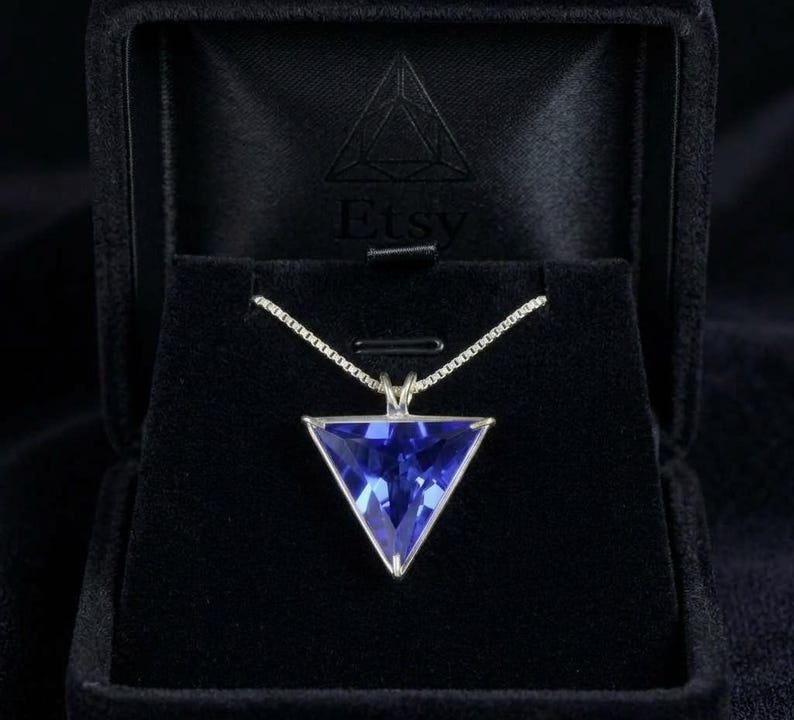 以下が含まれることがあります： 三角形の青い宝石のペンダントが付いたシルバーネックレス。ペンダントはシルバーのフレームにセットされ、繊細なチェーンから吊り下げられています。ネックレスは黒いジュエリーボックスに入っています。