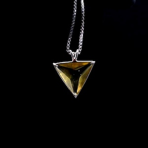 Puede incluir: Collar de plata con colgante triangular. El colgante tiene una gema facetada de color ámbar engastada en un marco de plata. La cadena es fina, tipo caja, sobre un fondo negro.
