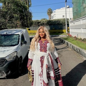 Puede incluir: Un vestido blanco con intrincados bordados burdeos y dorados. El vestido de manga larga presenta un cinturón envolvente y un borde estampado. El vestido se usa al aire libre en un día soleado.