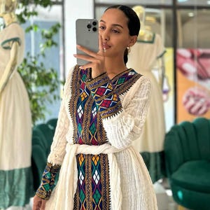 Puede incluir: Un vestido color crema con intrincados bordados en un vibrante patrón de rojo, azul, verde y amarillo. El vestido presenta mangas largas y un cinturón a juego. El bordado se concentra en el corpiño y los puños.