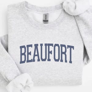 Puede incluir: Una sudadera gris claro con la palabra "BEAUFORT" impresa en azul marino en el pecho. La sudadera tiene cuello redondo y mangas largas con puños acanalados. La etiqueta de la marca Gildan es visible.