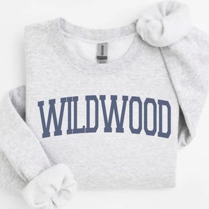 May include: A light gray sweatshirt with the word "WILDWOOD" printed in navy blue block letters across the front. The sleeves are cuffed, and the Gildan tag is visible at the neckline. The sweatshirt is made of a soft material.