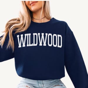 May include: A navy blue sweatshirt with the word 'WILDWOOD' printed in large, white, block letters across the chest. The sweatshirt has a crew neck and long sleeves. The person wearing the sweatshirt is also wearing light blue jeans and a black choker necklace.