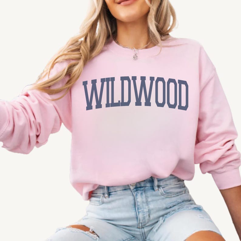May include: A light pink sweatshirt with the word "WILDWOOD" printed in navy blue block letters across the chest. The sweatshirt has long sleeves and a crew neck. The model is wearing light blue denim shorts.