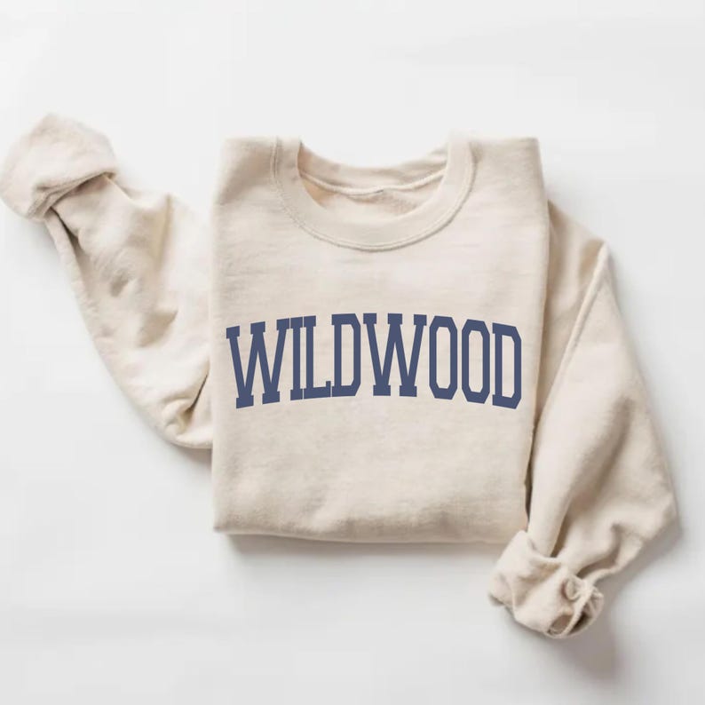 May include: A cream-colored sweatshirt with the word "WILDWOOD" printed in navy blue block letters across the chest. The sleeves are rolled up, and the sweatshirt is folded neatly. The fabric appears soft and comfortable.