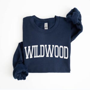 May include: A navy blue sweatshirt with the word "WILDWOOD" printed in large, white, block letters across the chest. The sweatshirt has a crew neck and long sleeves. The garment is displayed on a white background.