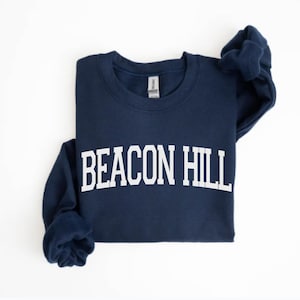May include: A navy blue sweatshirt with the text "BEACON HILL" in bold white letters. The sweatshirt is folded, with the sleeves draped to the side. The garment is made of a soft material.