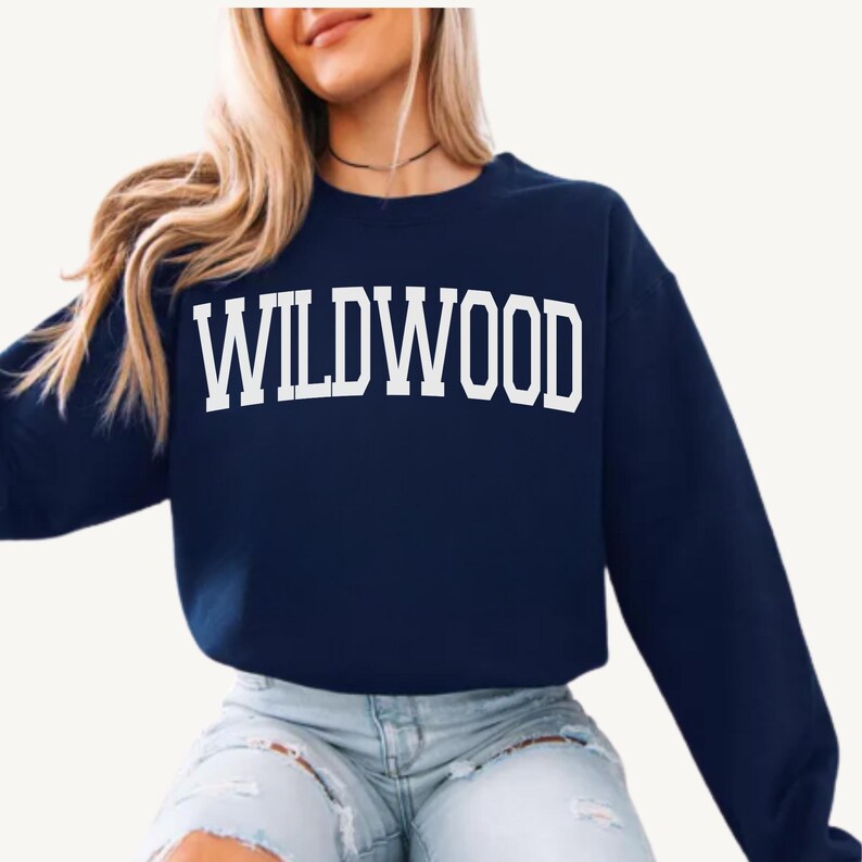 May include: A navy blue sweatshirt with the word 'WILDWOOD' printed in large, white, block letters across the chest. The sweatshirt has a crew neck and long sleeves. The person wearing the sweatshirt is also wearing light blue jeans and a black choker necklace.