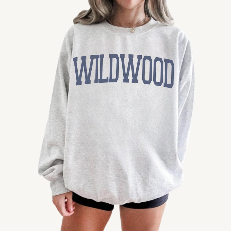 May include: A light gray sweatshirt with the word "WILDWOOD" printed in navy blue block letters across the chest. The sweatshirt has long sleeves and a crew neck.