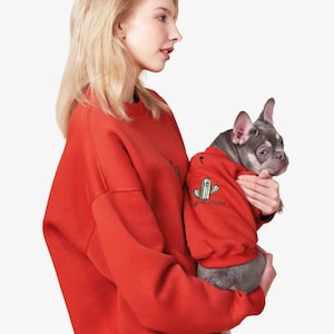 May include: A matching red sweatshirt and dog sweater set. The dog sweater features a cactus graphic. The woman is holding a dog wearing a matching red sweater with a cactus design. The set is a fun and stylish outfit for pet owners.