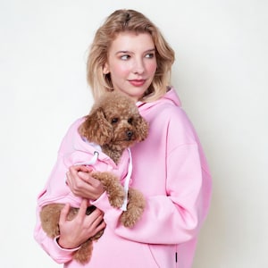 Puede incluir: Una mujer sostiene un perro pequeño marrón que lleva una sudadera con capucha rosa. El perro está acurrucado en los brazos de la mujer, que también lleva una sudadera con capucha rosa a juego. Ambas sudaderas tienen cordones blancos. La mujer lleva una sudadera con capucha rosa.
