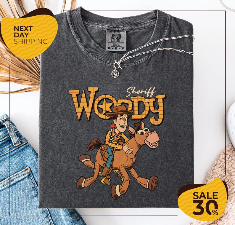 A gray t-shirt with a cartoon image of Woody, a character from the Toy Story franchise, and a sheriff badge on the neck. The t-shirt is displayed on a white background, and there are jeans and a sweater visible in the background.