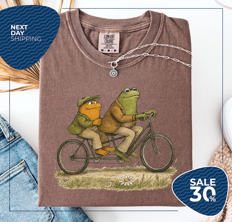 a brown t-shirt with a cartoon illustration of two frogs riding a bicycle, accompanied by a necklace and a sale sign indicating a 30% discount.