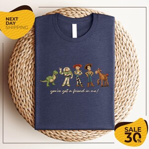 A blue t-shirt with a graphic design featuring characters from the Toy Story movie, including Woody, Buzz Lightyear, and a dinosaur. The text on the t-shirt says "you've got a friend in me!"