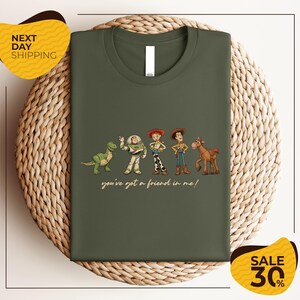 A green t-shirt with a graphic design featuring characters from the Toy Story movie, including Woody, Buzz Lightyear, and a dinosaur. The text below the characters reads "you've got a friend in me!"