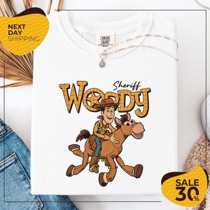 a white t-shirt with a cartoon image of Woody, a character from the Toy Story movie, riding a horse. The t-shirt is displayed on a white background, and there are yellow and black sale signs visible in the image.