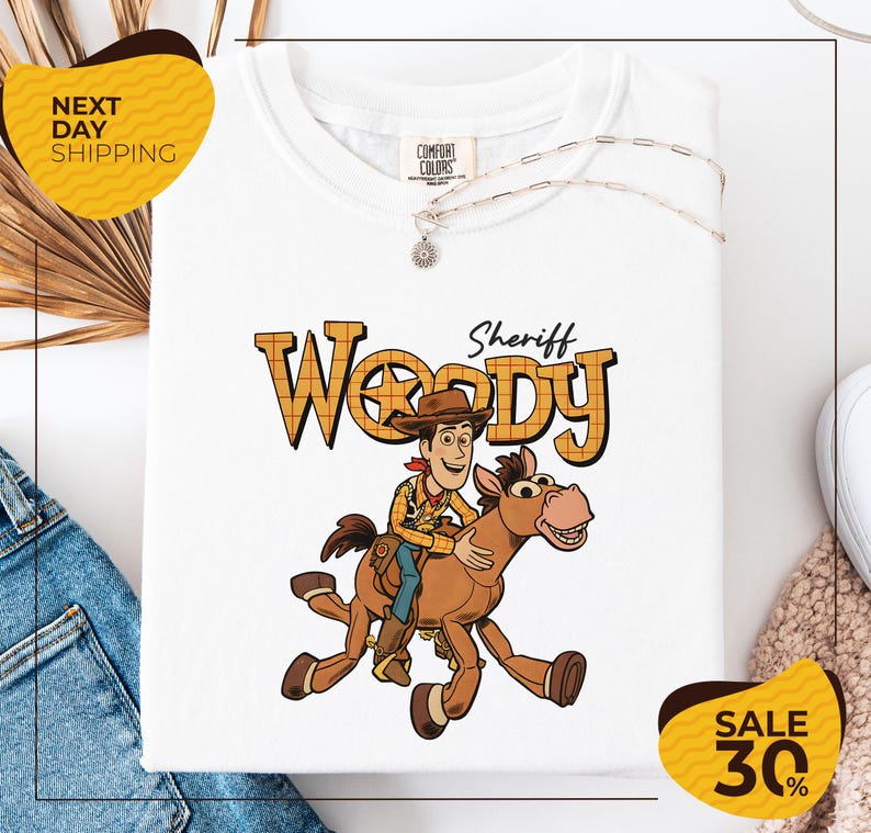 a white t-shirt with a cartoon image of Woody, a character from the Toy Story movie, riding a horse. The t-shirt is displayed on a white background, and there are yellow and black sale signs visible in the image.