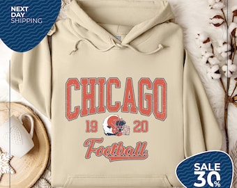 May include: A beige hoodie with a distressed orange graphic that reads "CHICAGO" above "19 20 Football". A football helmet graphic is in the center. The hoodie has a drawstring hood and a front pocket. The text "NEXT DAY SHIPPING" is in a blue oval.