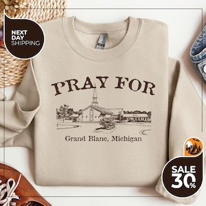May include: Beige crewneck sweatshirt with the text "PRAY FOR" above a church illustration. Below the illustration is "Grand Blanc, Michigan". The sweatshirt has a classic fit and long sleeves.