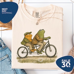 a white t-shirt with a drawing of two frogs riding a bicycle, along with a tag that reads "COMFORT COLLECTIONS". The t-shirt is displayed on a white background, and there is a sale sign indicating a 30% discount.