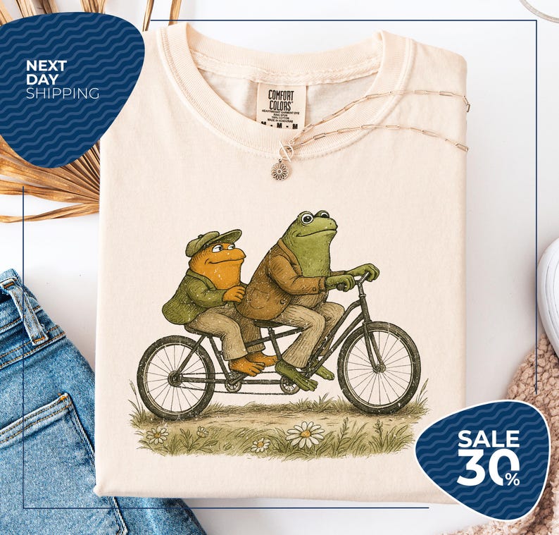 a white t-shirt with a drawing of two frogs riding a bicycle, along with a tag that reads "COMFORT COLLECTIONS". The t-shirt is displayed on a white background, and there is a sale sign indicating a 30% discount.